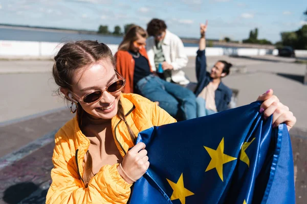 Mujer feliz en auriculares y gafas de sol sosteniendo la bandera de la UE cerca de amigos borrosos - foto de stock