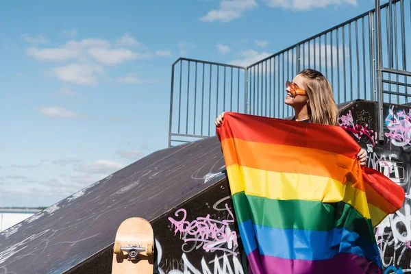 Heureuse jeune femme tenant drapeau lgbt près de rampe de skate et planche à roulettes — Photo de stock