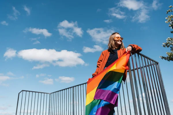 Baixo ângulo de visão da mulher sorridente com bandeira lgbt contra o céu azul nublado — Fotografia de Stock