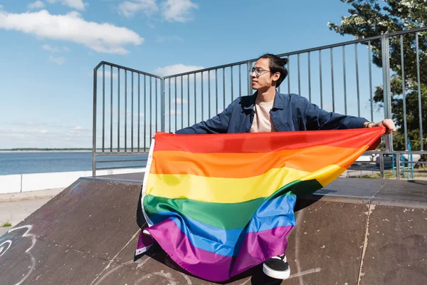Joven asiático hombre con lgbt orgullo bandera sentado en rampa en skate park - foto de stock