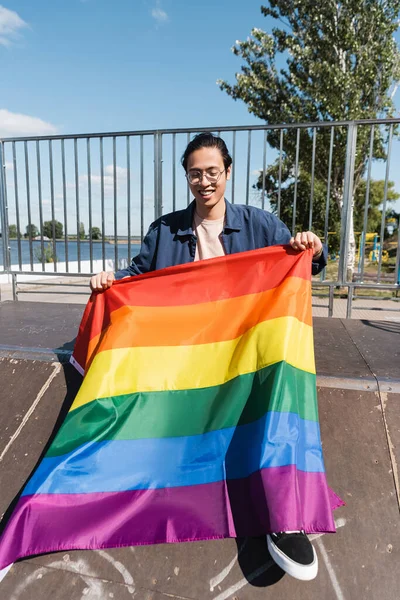 Счастливый азиатский мужчина в очках сидит на рампе в скейт-парке с флагом — стоковое фото