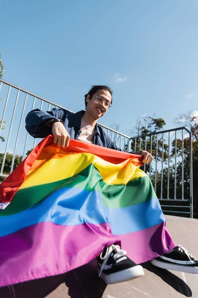 Low angle view of young asian man sitting with lgbt flag outdoors — Stock Photo