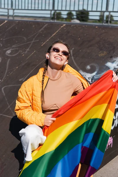 Femme heureuse dans les écouteurs sans fil assis avec drapeau lgbt sur la rampe dans le skate park — Photo de stock