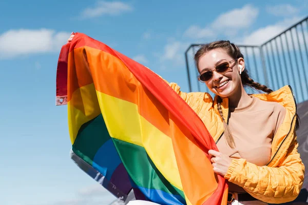 Cheerful woman in stylish sunglasses holding lgbt flag against blue sky — Stock Photo