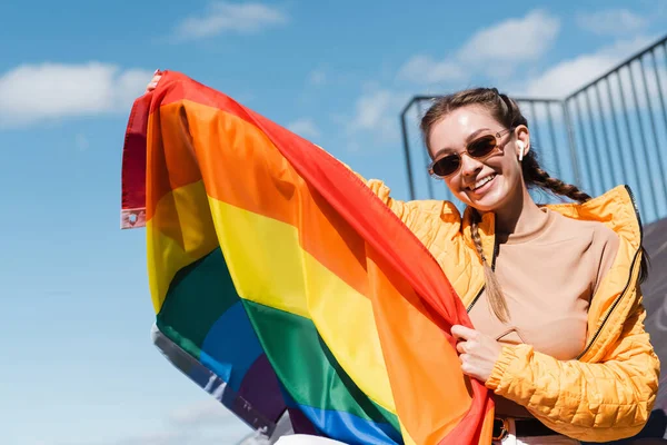 Mulher alegre em óculos de sol elegantes segurando bandeira lgbt contra o céu azul — Fotografia de Stock