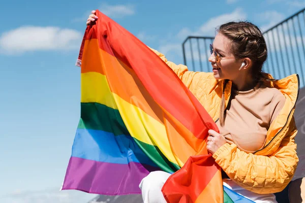 Smiling woman in earphone holding lgbt pride flag outdoors — Stock Photo