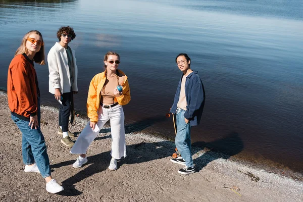 Trendy multiethnic friends standing near lake and looking at camera — Stock Photo