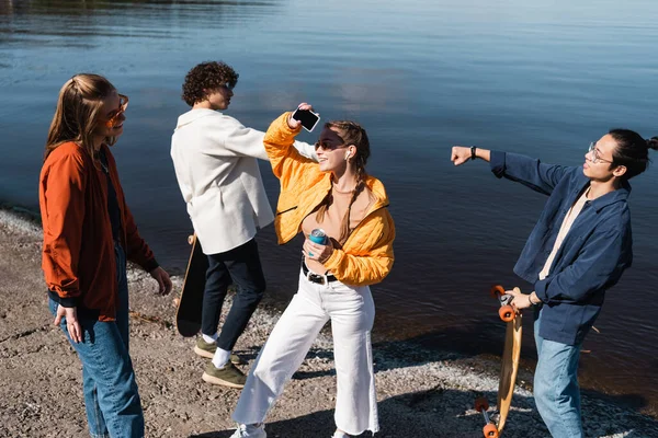 Donna sorridente con lattina di soda e smartphone vicino a pattinatori multietnici sulla riva del fiume — Foto stock