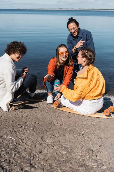 Happy multicultural skaters with smartphones talking near river — Stock Photo