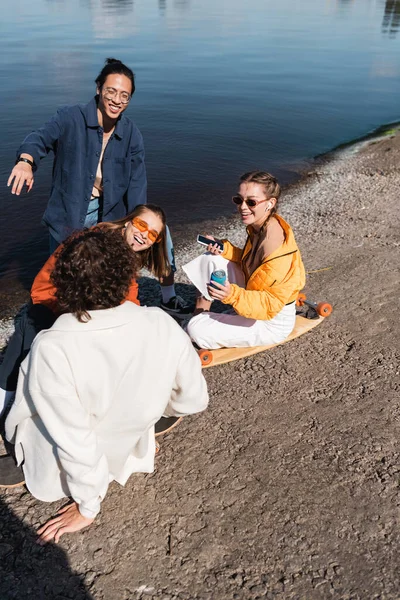 Happy woman with smartphone and soda can sitting on longboard near multiethnic friends — Stock Photo