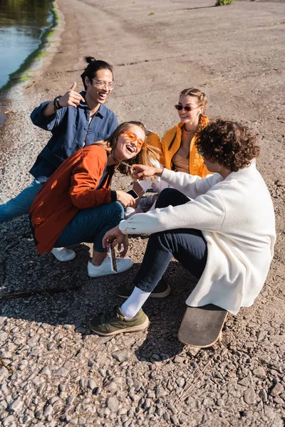 Alegre asiático hombre mostrando pulgar arriba cerca amigos tener divertido en río orilla - foto de stock