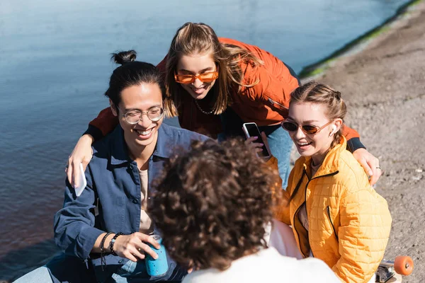 Mujer alegre abrazando amigos multiétnicos cerca del río - foto de stock