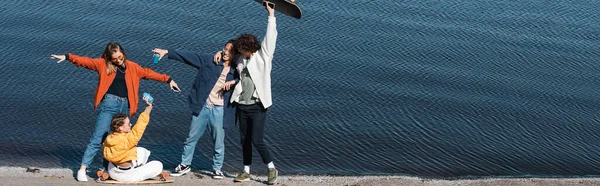 Happy multiethnic skaters in stylish clothes having fun near river, banner — Stock Photo