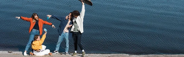 Patinadores multiétnicos felizes em roupas elegantes se divertindo perto do rio, bandeira — Fotografia de Stock