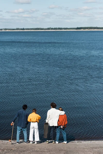 Vista trasera de mujeres jóvenes de pie cerca de patinadores en la orilla del río - foto de stock