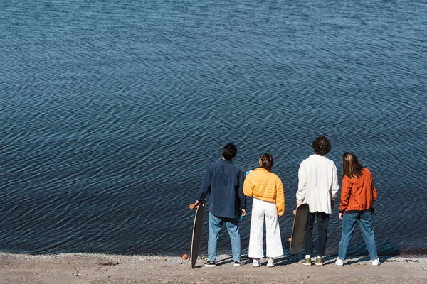 Vue arrière de jeunes patineurs en vêtements tendance debout près de la rivière — Photo de stock