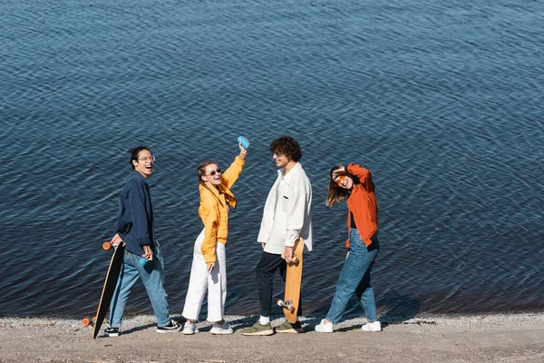 Pleine longueur de femme heureuse avec canette de soda dans la main levée près d'amis multiethniques sur la rive de la rivière — Photo de stock