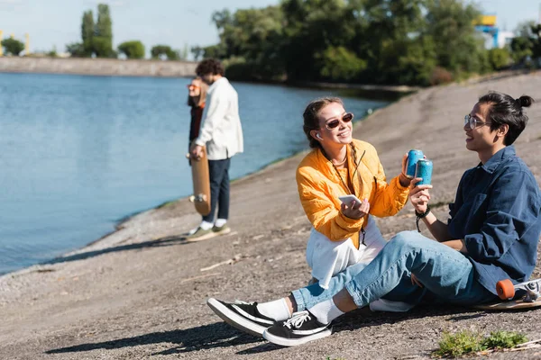 Asian man sitting on longboard near river and clinking soda cans with cheerful friend — Stock Photo