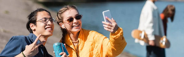 Glückliche Frau mit Sonnenbrille macht Selfie mit asiatischem Freund und zeigt Siegesgeste, Banner — Stockfoto