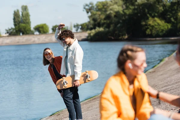 Femme gaie dans des lunettes de soleil souriant ami proche avec planche à roulettes sur le bord de la rivière — Photo de stock