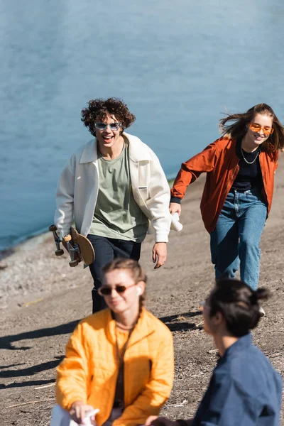 Uomo allegro con skateboard in esecuzione sulla riva del fiume vicino agli amici multiculturali — Foto stock