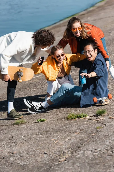 Patinadores multiculturais alegres com lata de refrigerante e smartphone se divertindo no dique — Fotografia de Stock