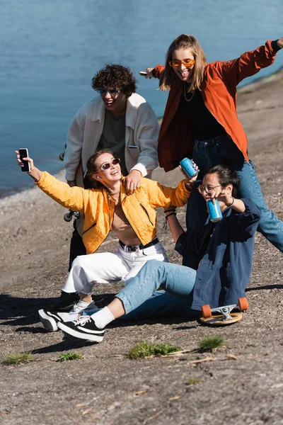 Femme heureuse avec smartphone et soda peut s'amuser avec des amis interracial sur le remblai — Photo de stock