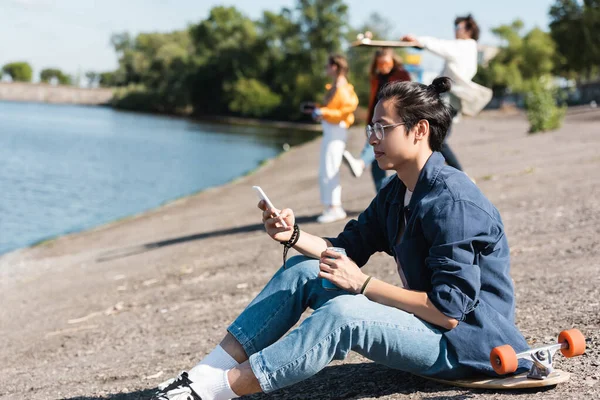 Asiatique l'homme dans les lunettes à l'aide de téléphone mobile tandis que assis sur longboard près de rivière et flous amis — Photo de stock