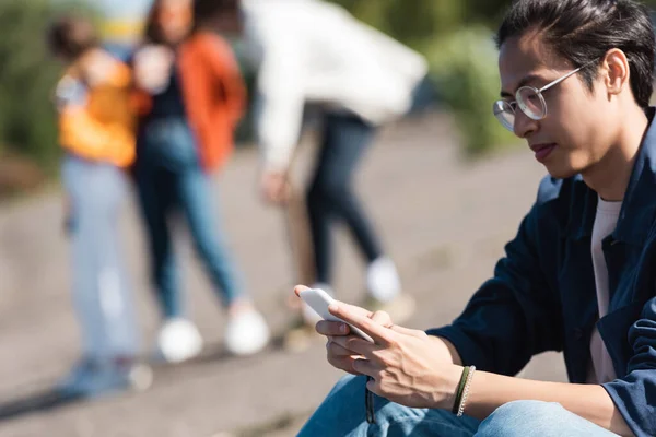 Asiático hombre en gafas usando móvil cerca amigos en borrosa fondo - foto de stock