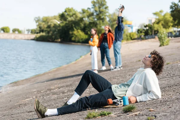 Full length of young skater relaxing on embankment near blurred friends — Stock Photo