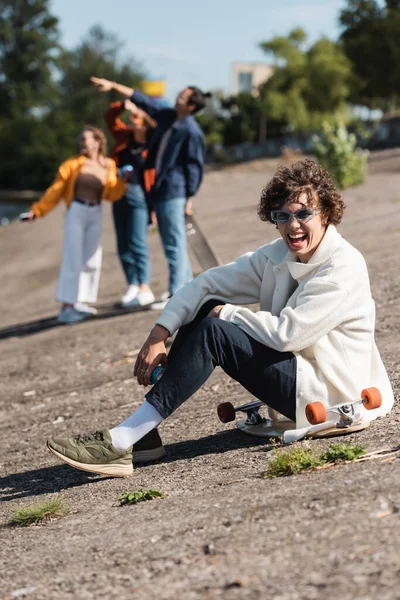 Homem animado em óculos de sol sentado no longboard e rindo da câmera ao ar livre — Fotografia de Stock