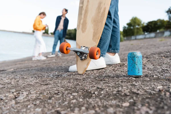 Vista parziale del pattinatore con longboard vicino alla lattina di soda e gli amici offuscati sulla riva del fiume — Foto stock