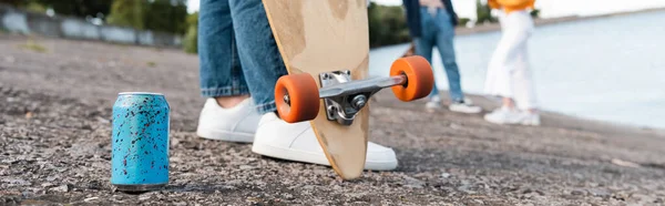 Vista parcial de la mujer con longboard cerca de lata de refresco en asfalto y amigos borrosos, bandera - foto de stock