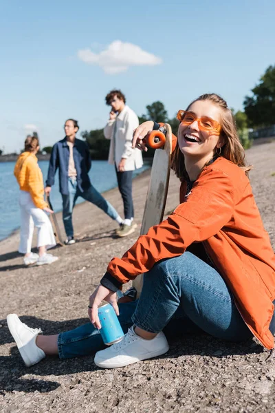 Laughing woman with penny board looking at camera while sitting on river bank near blurred friends — Stock Photo