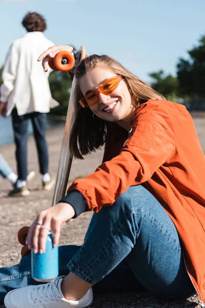 Fröhliche Frau mit Sonnenbrille blickt in die Kamera neben Skateboard und Getränkedose — Stockfoto