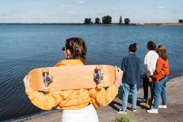 Vista posterior de la mujer joven con monopatín de pie cerca del río y amigos borrosos - foto de stock