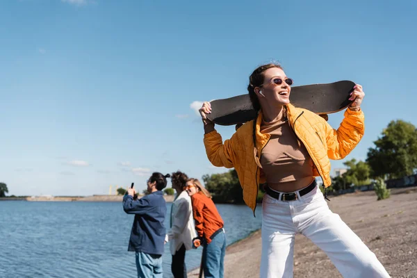 Femme excitée avec planche à roulettes debout près des amis flous sur le bord de la rivière — Photo de stock