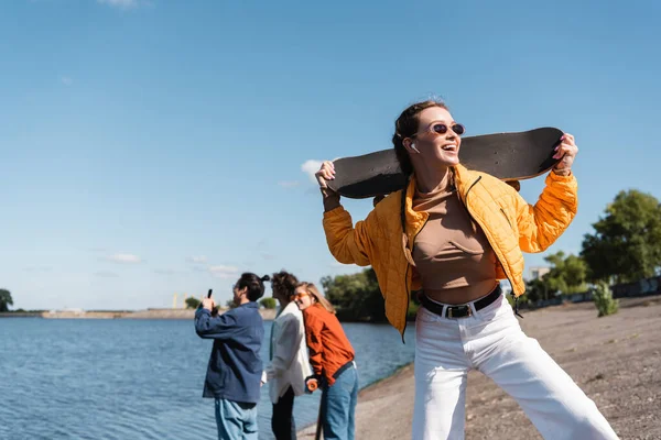 Donna eccitata con skateboard in piedi vicino a amici offuscati sul lungofiume — Stock Photo