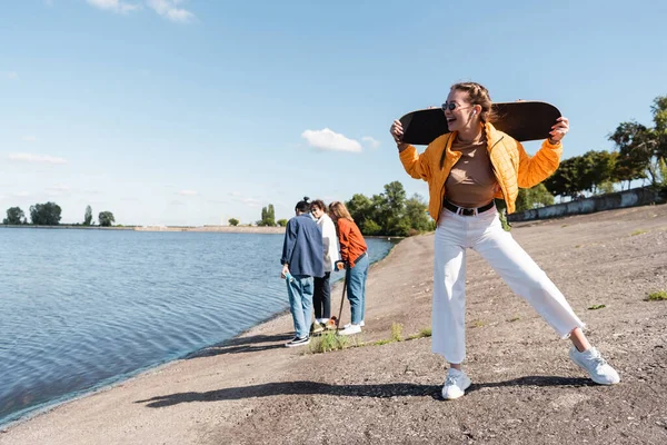 В полный рост взволнованная женщина с скейтбордом, стоящая на берегу реки рядом с друзьями на заднем плане — стоковое фото