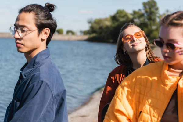 Mujer complacida en gafas de sol sonriendo con los ojos cerrados cerca del río y borrosa amigos interracial - foto de stock