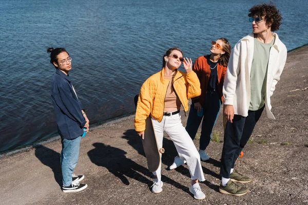 Young woman with skateboard adjusting sunglasses near interracial friends on river bank — Stock Photo