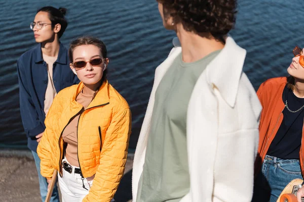 Mujer joven en gafas de sol de moda mirando a la cámara cerca de amigos interracial - foto de stock