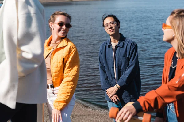 Young asian man looking at camera near happy friends on riverside — Stock Photo