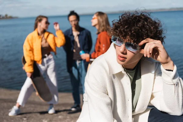 Bouclé homme réglage des lunettes de soleil près des amis flous sur le bord de la rivière — Photo de stock