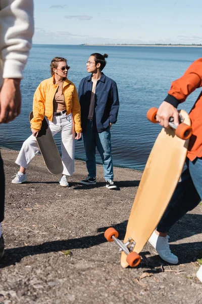 Stylish interracial skaters standing on riverside near blurred friends — Stock Photo