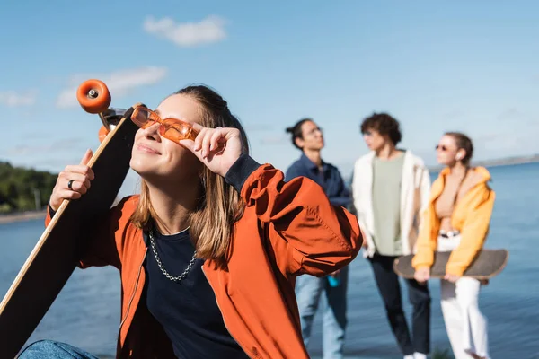 Pleased woman with longboard adjusting trendy sunglasses near friends on blurred background — Stock Photo