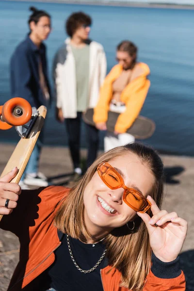 Femme gaie avec penny planche réglage des lunettes de soleil près des amis flous — Photo de stock