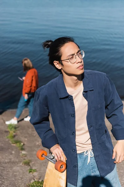 Young asian skater in blue shirt and eyeglasses looking away on riverside — Stock Photo