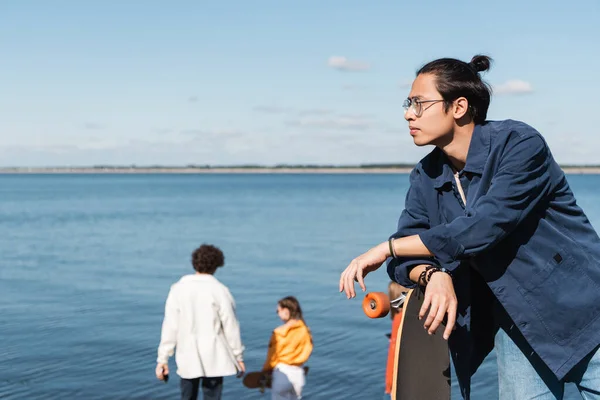 Jovem asiático skater olhando afastado perto desfocado amigos e rio — Fotografia de Stock