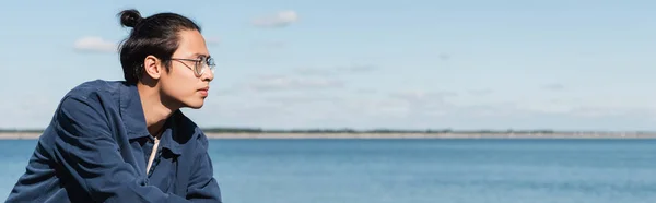 Young asian man in blue shirt and eyeglasses looking away near river, banner — Stock Photo